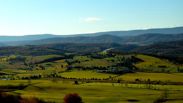 Rolling green fields in the foreground, with forested mountains topped with windmills in the distance