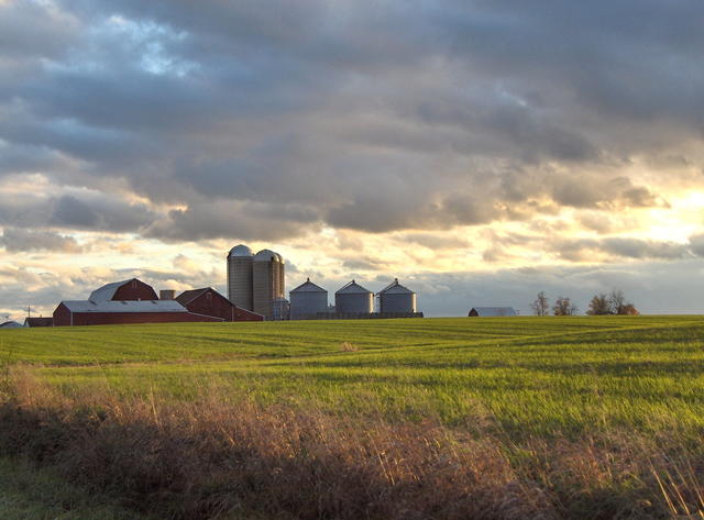 A farm with silos on the horizon, cropland in the foreground, under a cloudy sky with late afternoon sun shining through