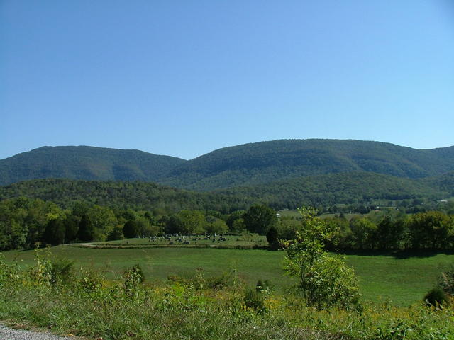 A flat, grassy valley with forested hills in the background, and steep mountains behind that.