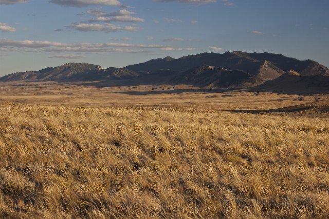 Flat, dry grasslands in the foreground with low mountains in the background, under a blue sky with some small clouds