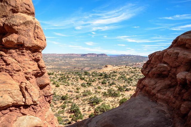 Reddish rock formations on either side, opening to a desert with scattered small scrubs, tablelands in the distance