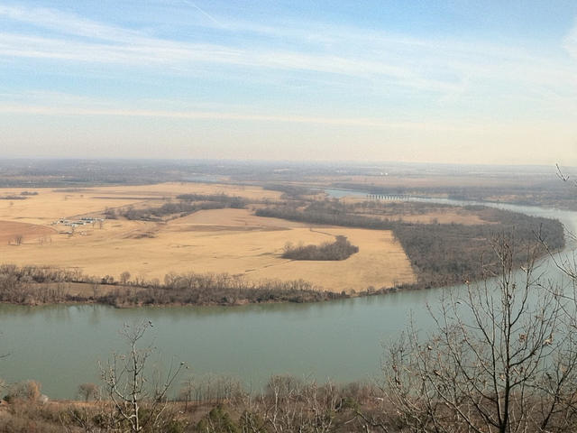 A large river gently curving around a large, flat area of land, partly used for agriculture, and with some forest.