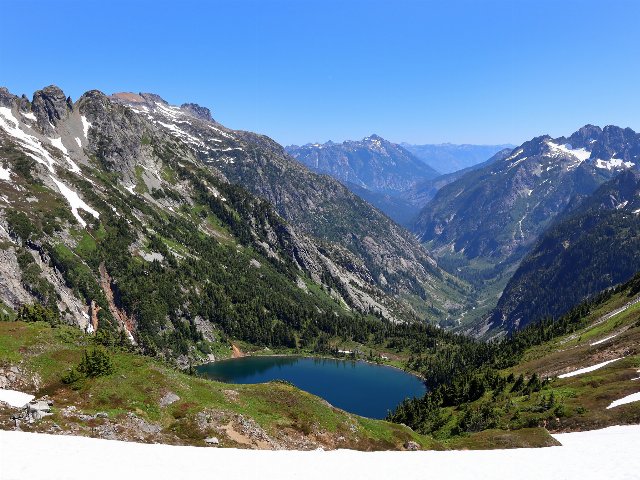 Snow in the foreground, a small lake, some coniferous forest, and rugged snow-capped mountains with a U-shaped valley.