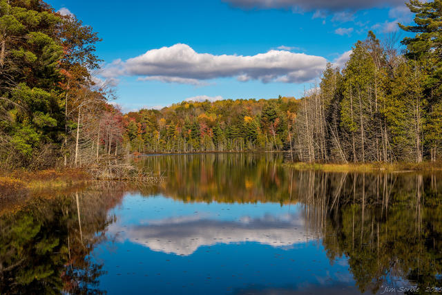 lake with still, reflective water, forest of conifers and hardwoods changing color on the far shore
