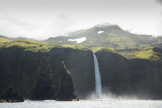 waterfall over a steep cliff into the ocean, in front of a greenish mountain disappearing into clouds and fog