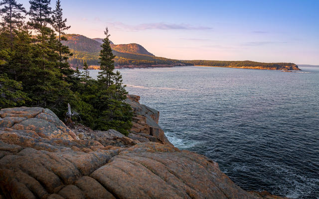 rocky coastline with conifers, ocean with fine waves, and rounded mountains in distance