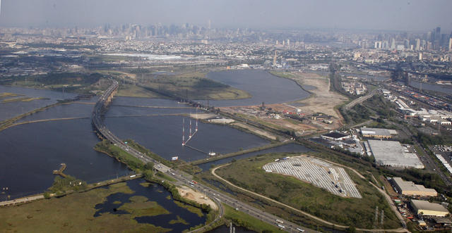 A road crossing a tidal river, with marshes and industrial development on either side, a big city skyline in the background.